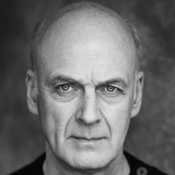 William Chubb wearing a dark suit and glasses, smiling against a plain grey background.