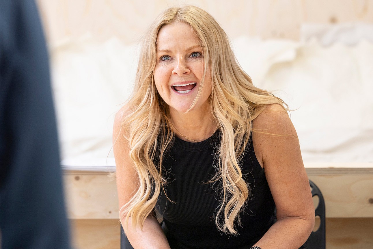 A woman with long blonde hair smiles broadly, wearing a black top, in a rehearsal setting.