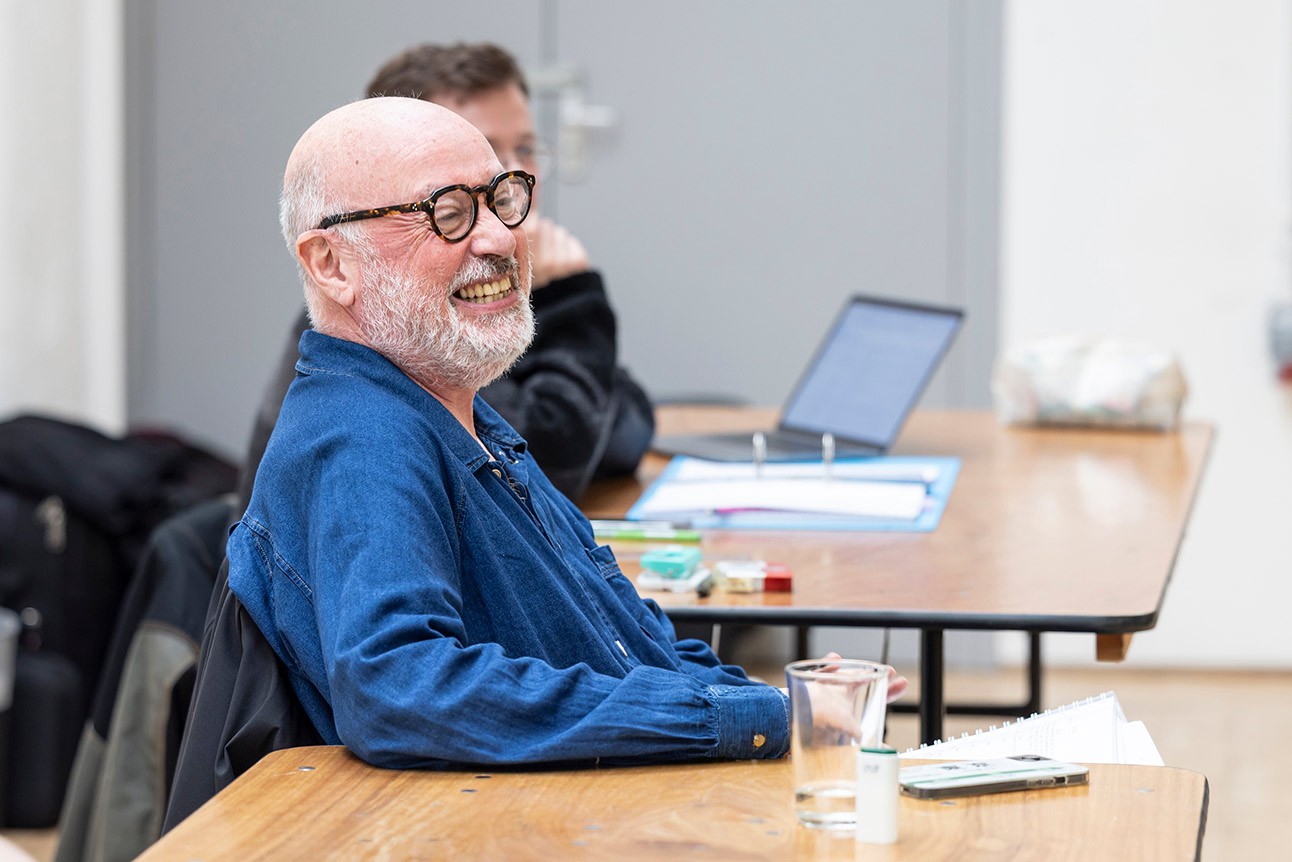 A man in glasses and blue shirt smiling, seated at a wooden table with papers.
