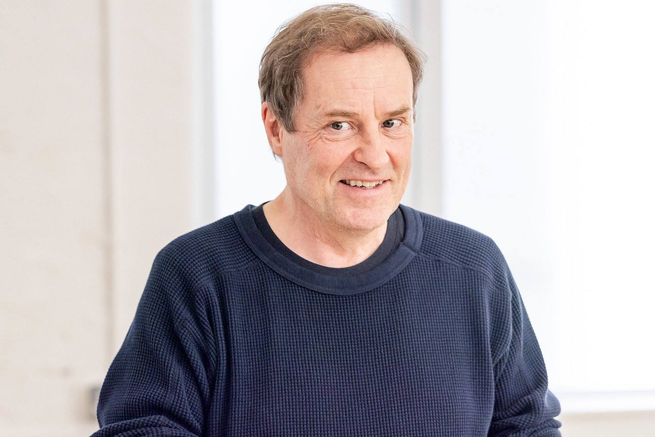 A man in a navy jumper smiles, standing in a bright room with white walls.