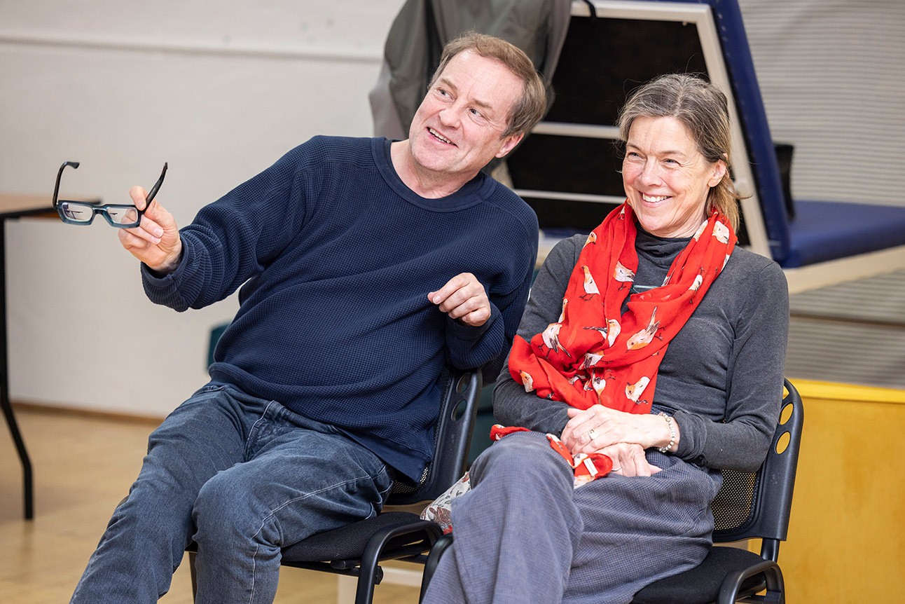 Two people seated, smiling during rehearsals; man in navy jumper, woman in red scarf.