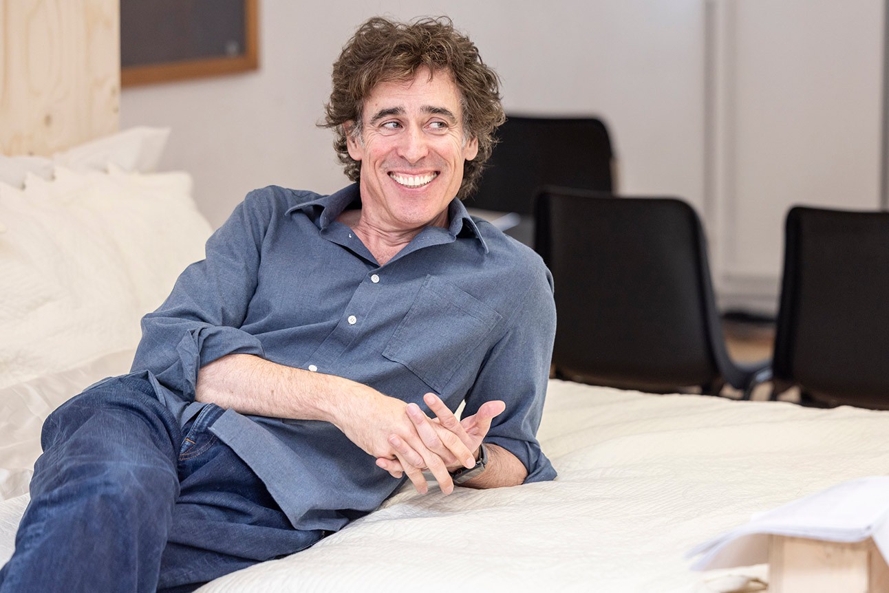 Man in blue shirt smiling, reclining on a white surface, with chairs in the background.