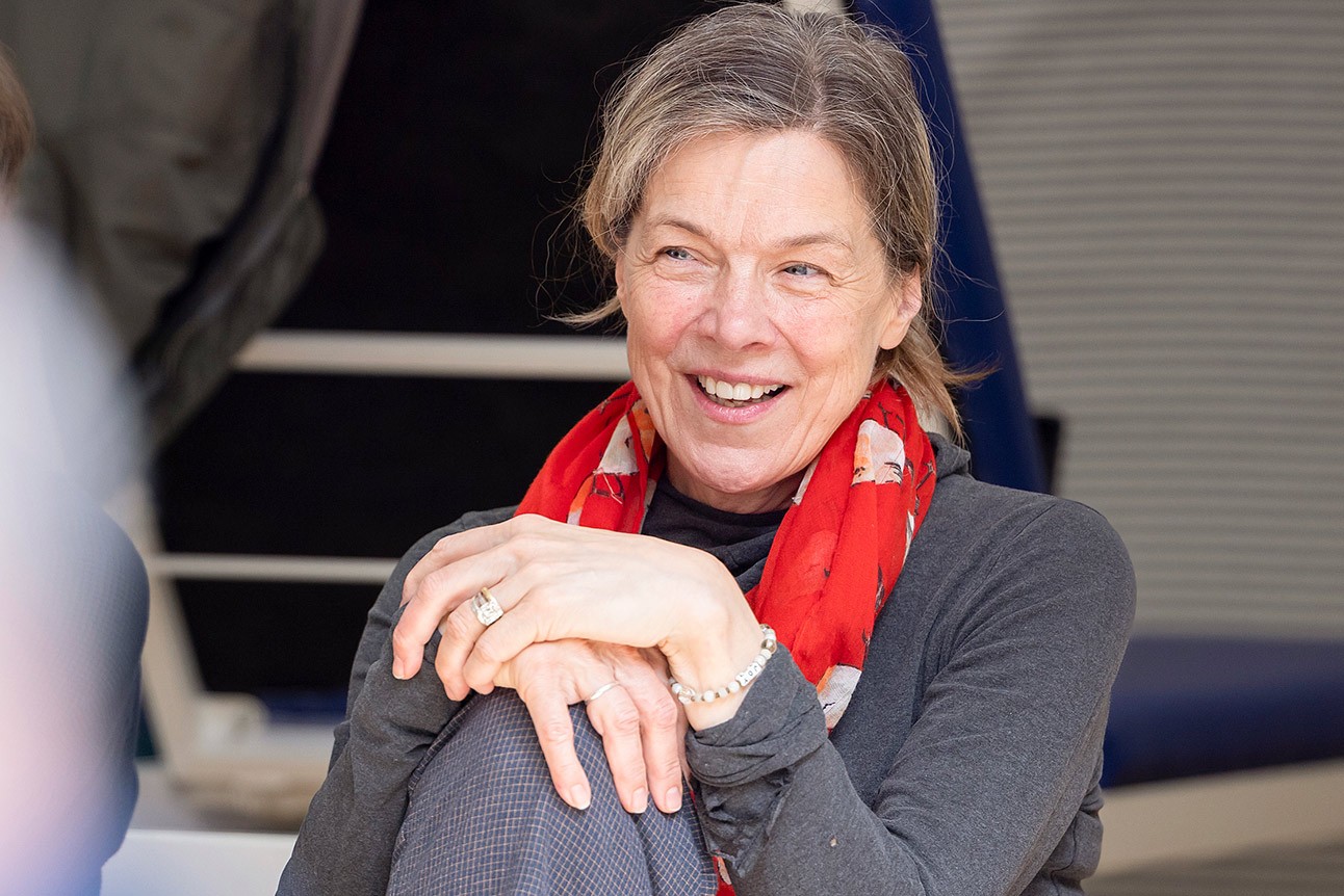Smiling woman with grey hair and red scarf, seated with hands clasped, in rehearsal setting.