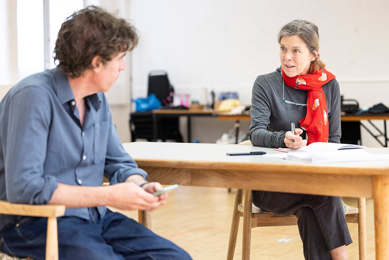 Two people in discussion at a table, woman wearing a red scarf, rehearsal setting.
