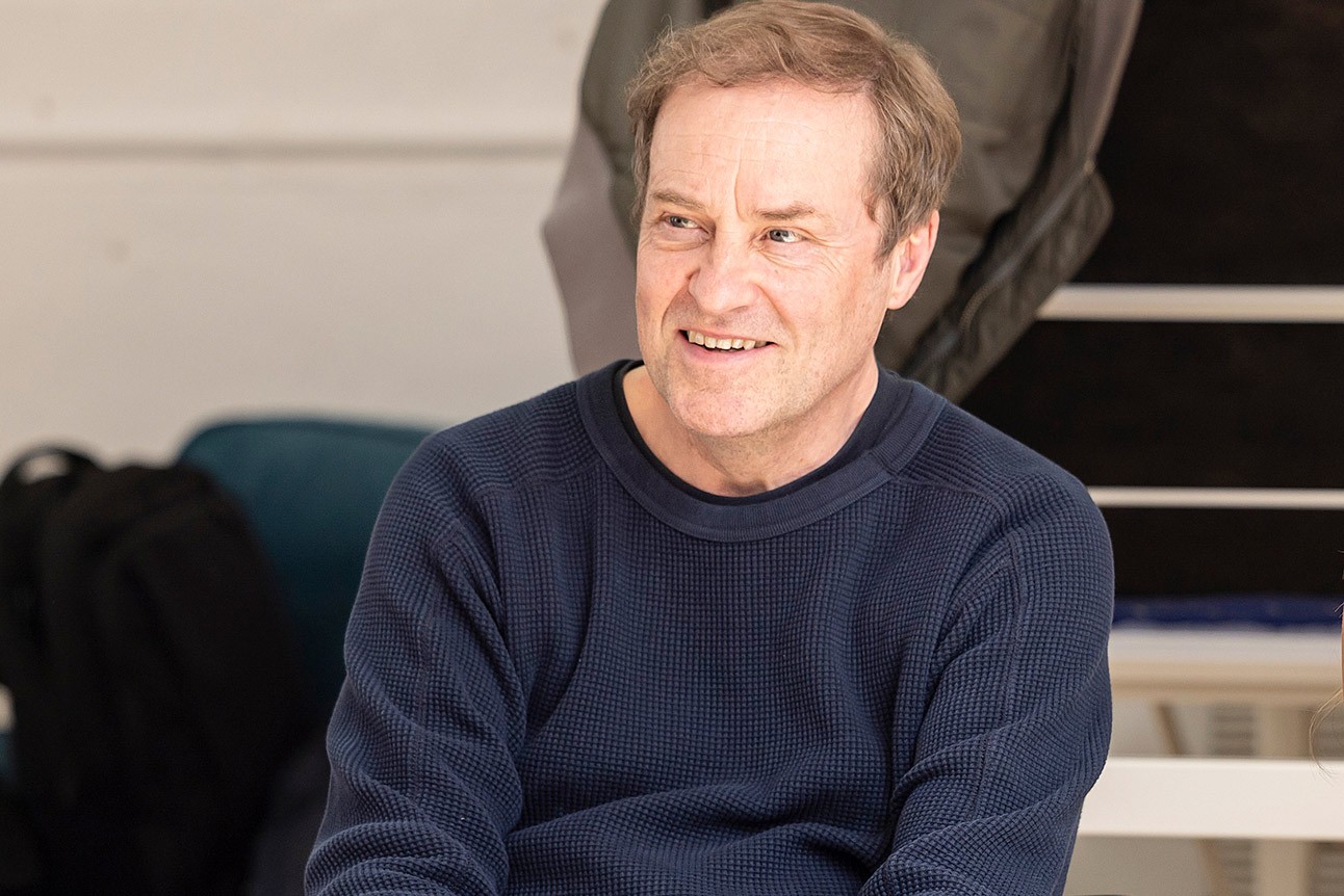 Smiling man in a navy jumper sits, light background, during "The Truth" rehearsals.