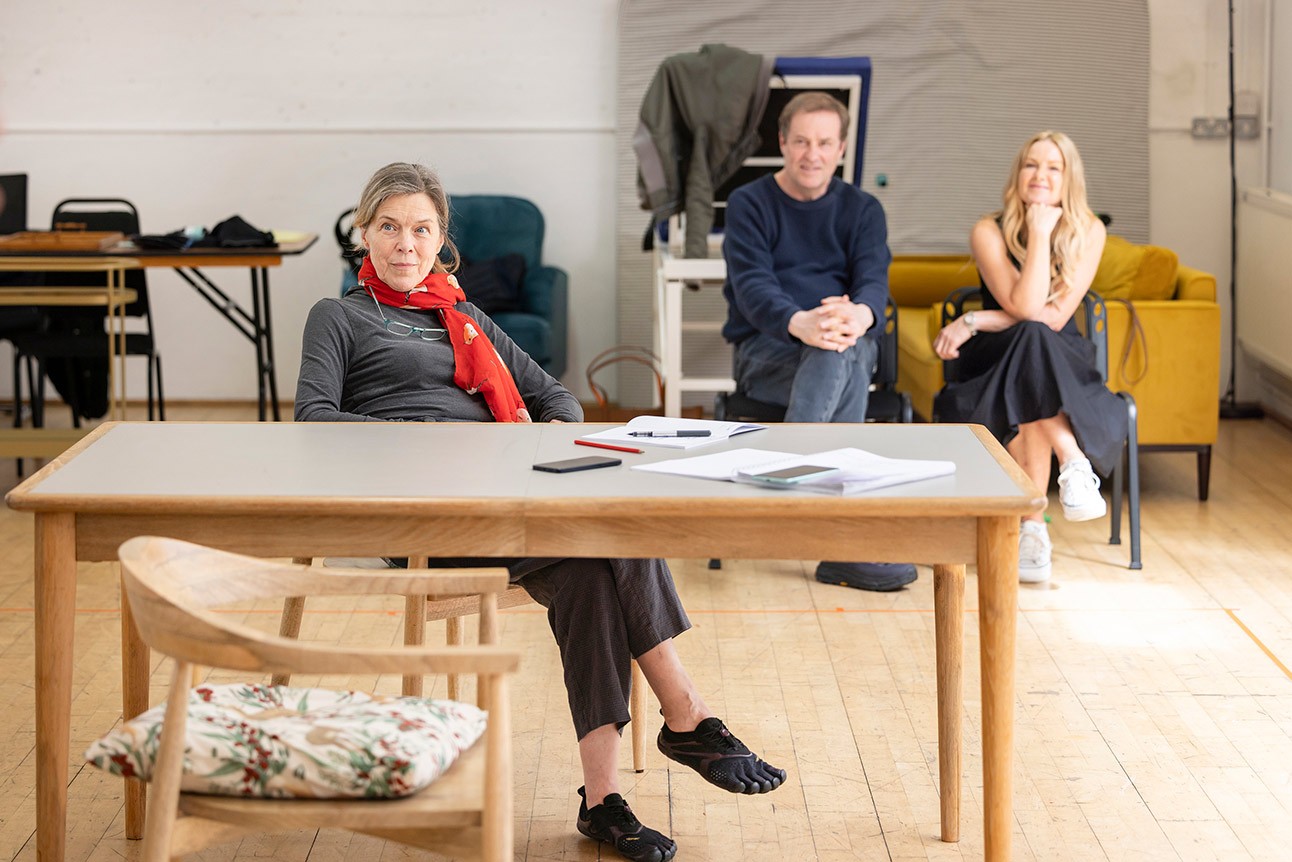 Three people seated in a rehearsal room, one woman in a red scarf at a table.