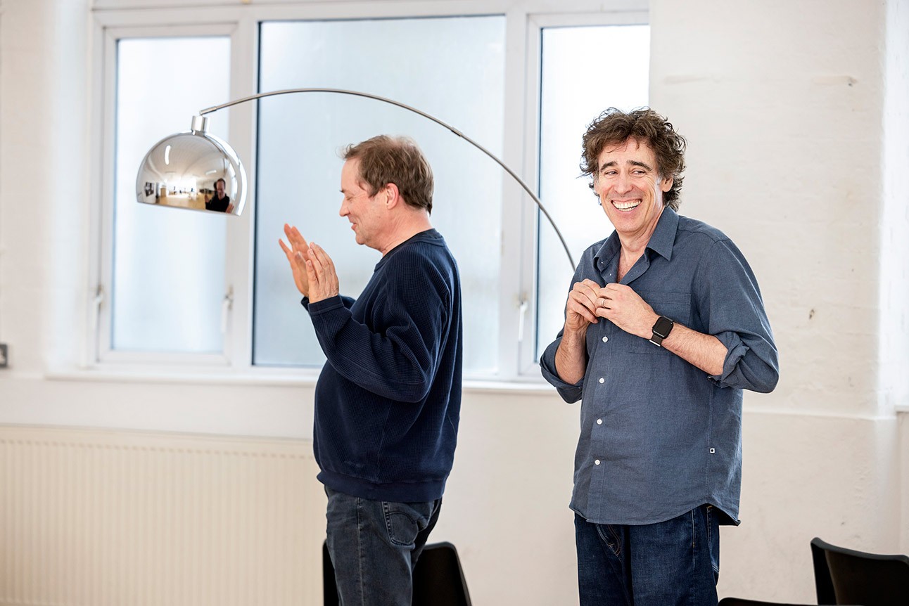Two men in casual clothing, one clapping, the other smiling, under a modern lamp.
