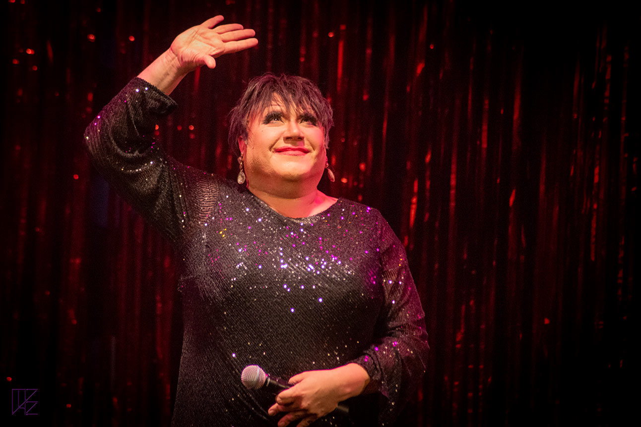 Trevor Ashley performs on stage, smiling, with a vibrant red curtain backdrop.