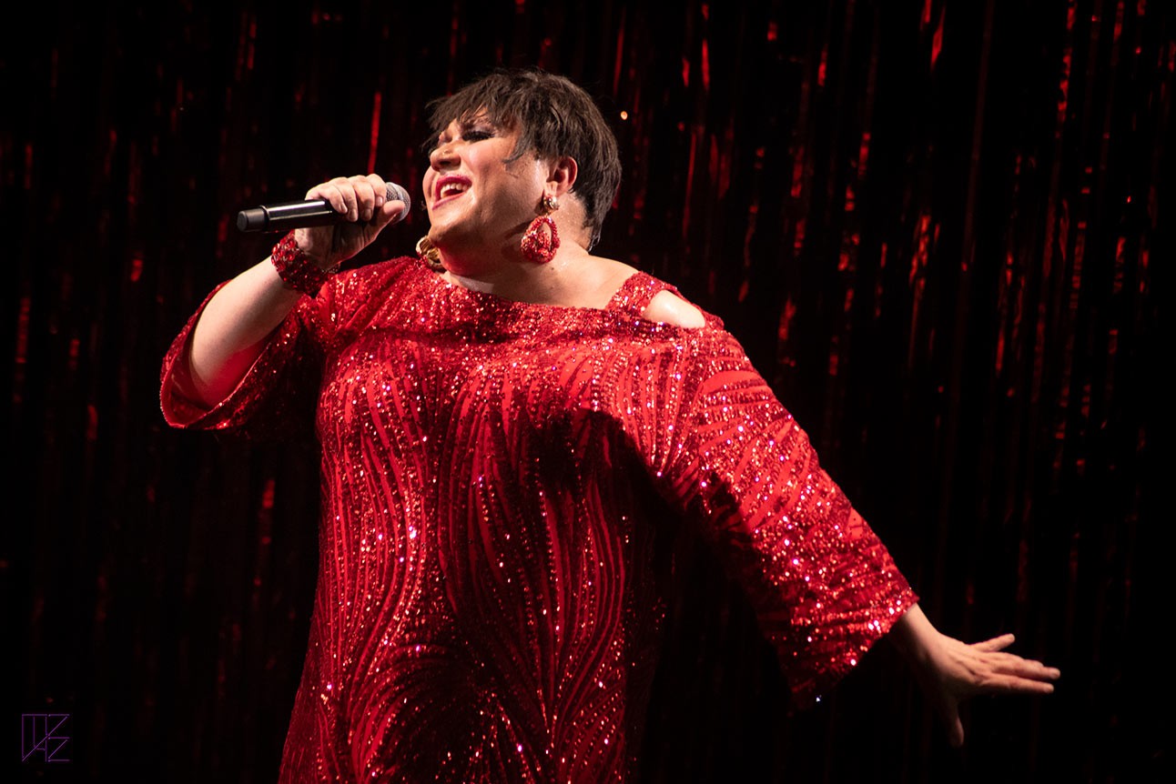 Trevor Ashley on stage, vibrant costumes, spotlight, black background.