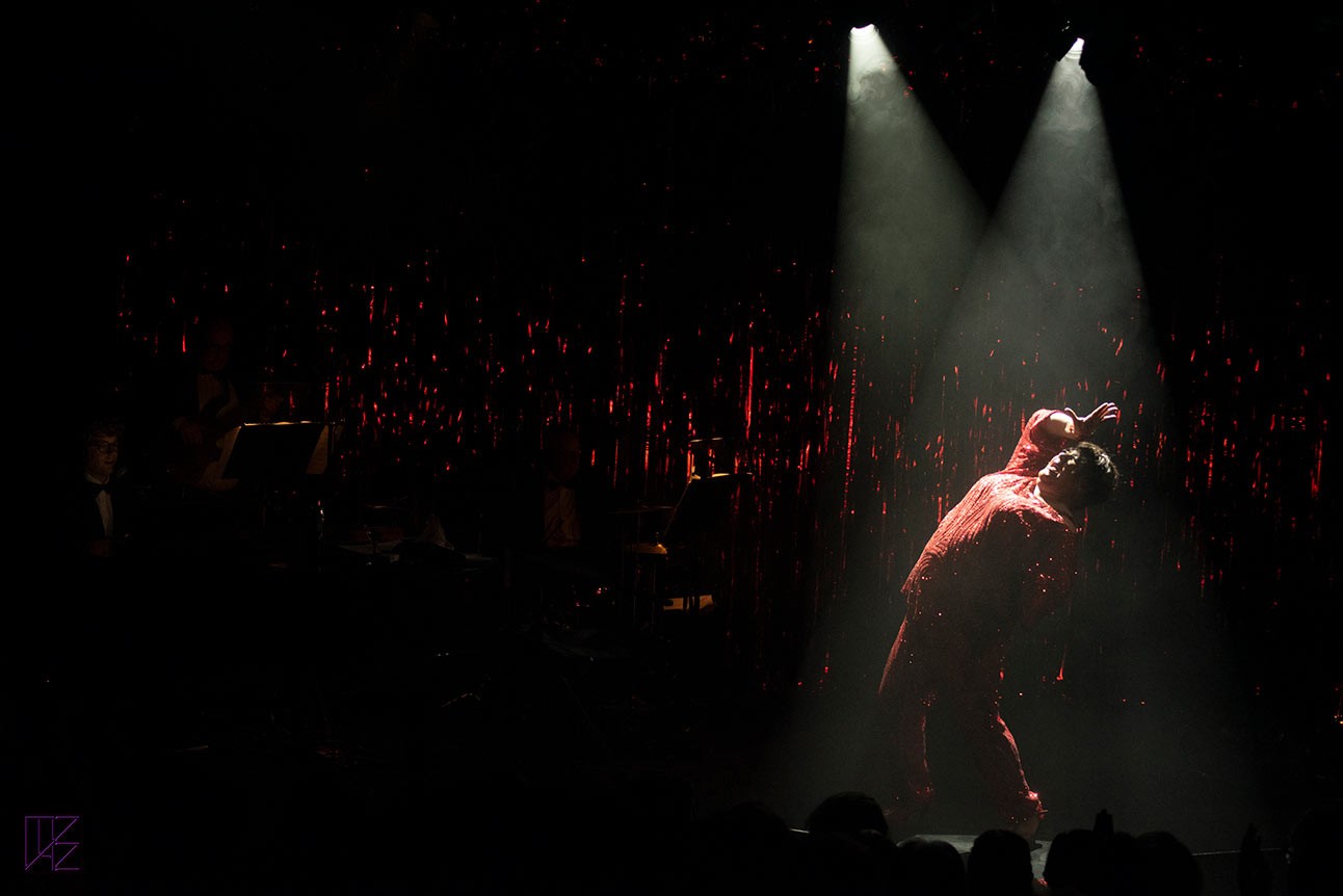 Performer in red sequined outfit poses dramatically under spotlight on dark stage with musicians.