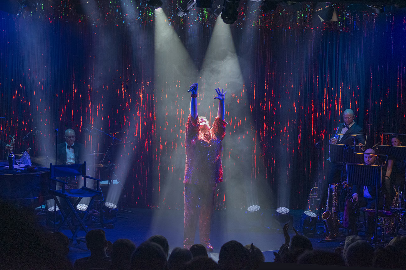 Trevor Ashley performs on stage in a sequined gown, holding a microphone, with a spotlight background.