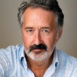 A man with grey hair and beard wearing a light blue shirt, neutral background.