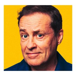 Ardal O'Hanlon smiling, wearing a dark shirt, standing against a plain yellow background.