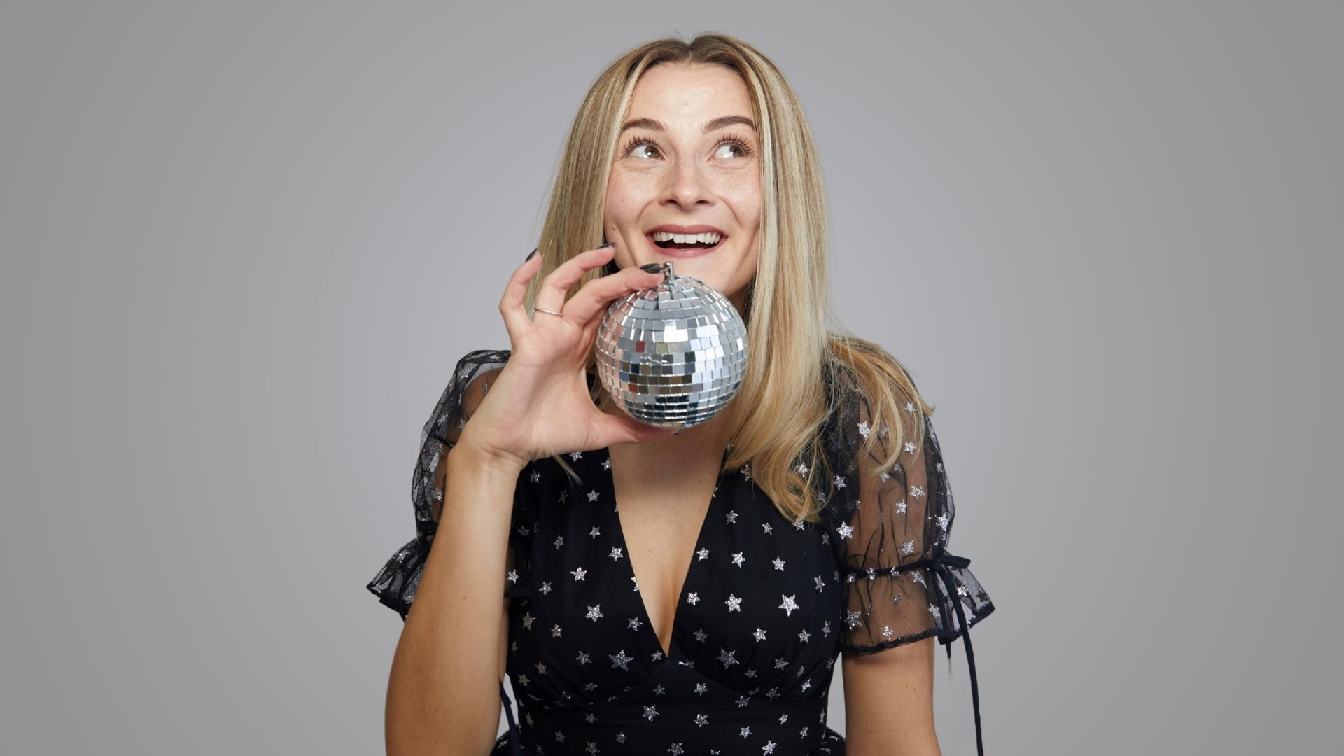 Hannah Lowther in star-patterned dress holds a small disco ball, smiling against a grey background.