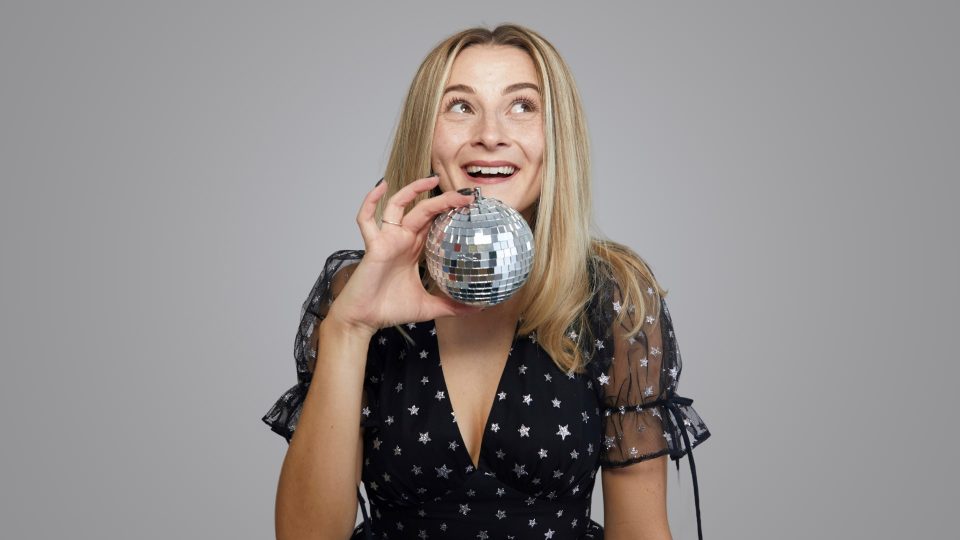 Hannah Lowther in star-patterned dress holds a small disco ball, smiling against a grey background.