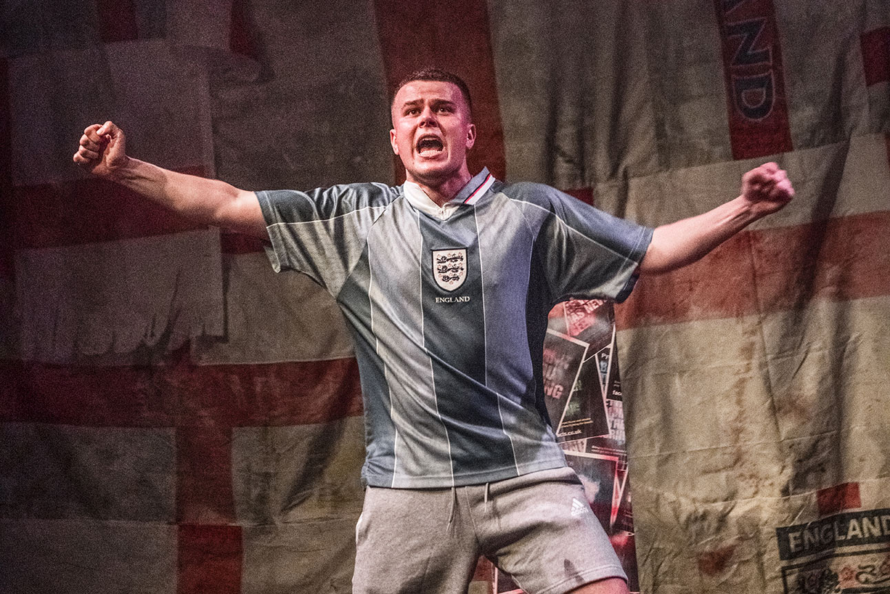 Man holding flare, wearing England football shirt, red smoke billowing, crowd cheering in background.