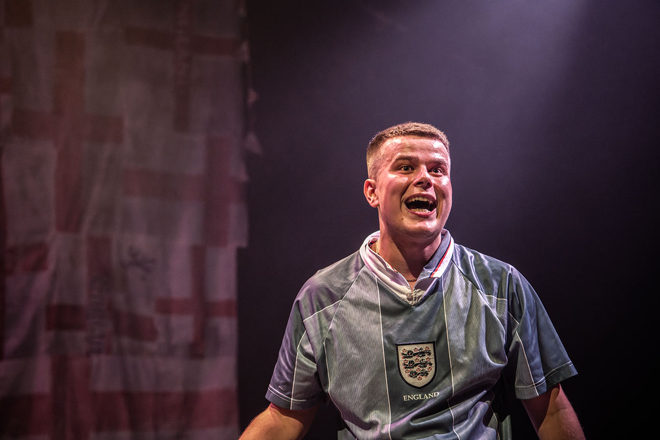 Man holding lit flare behind him, wearing England football shirt, surrounded by cheering crowd in stadium.