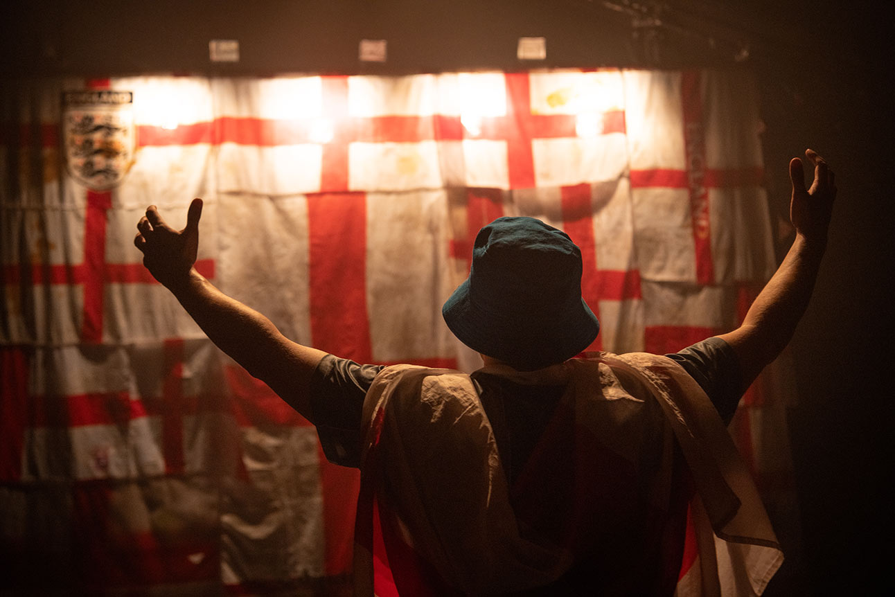 Man holding lit flare, smoke rising, wearing England football shirt, surrounded by cheering crowd, daytime.
