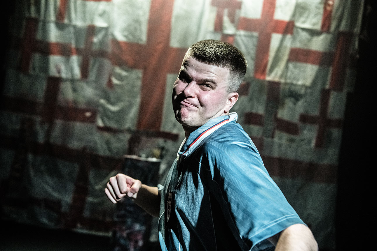 Man wearing England football shirt holds lit flare behind him, surrounded by cheering crowd in stadium.
