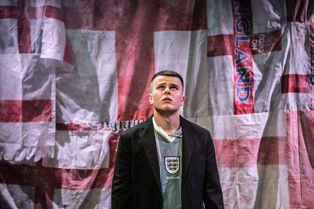 Man holding lit flare behind him, wearing white England football shirt, surrounded by cheering crowd.