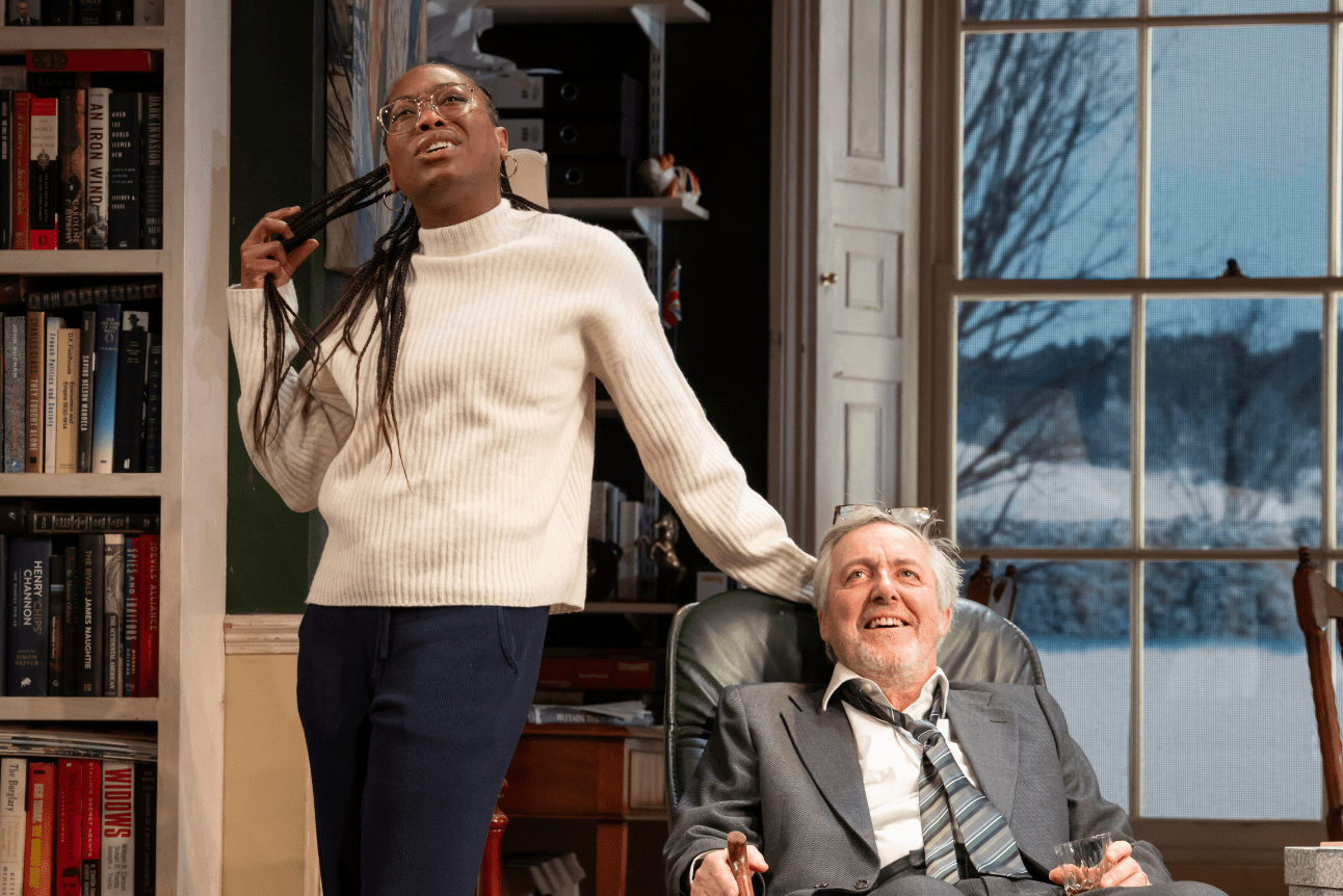 Sophie stands playing with her hair while talking. Jim Hacker is sat in a desk chair in front of her.