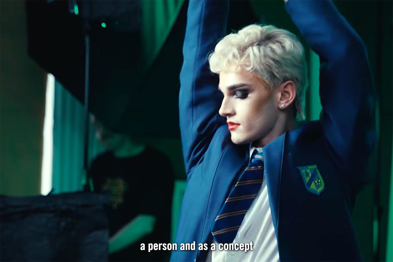 Person with short blonde hair in blue school uniform, arms raised, dramatic lighting, text overlay.