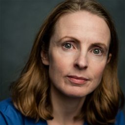 A woman with shoulder-length brown hair and a blue top, against a grey background.