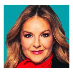 Sarah Hadland smiling, wearing a black top and red scarf, against a plain blue background.