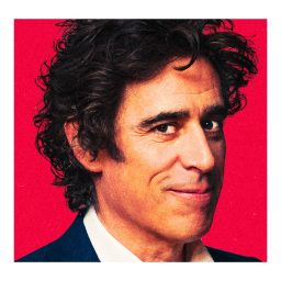 Stephen Mangan smiling, wearing a dark suit and white shirt, against a red background.