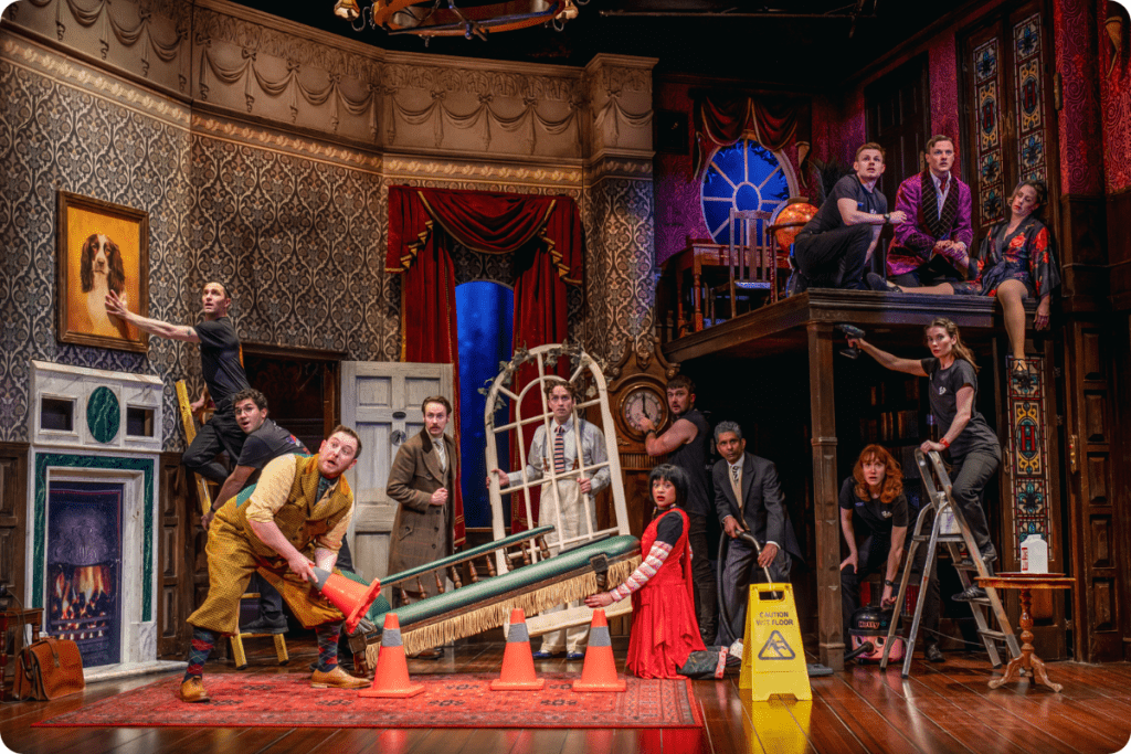 A chaotic theatre scene features actors in colourful costumes amidst a richly detailed, vintage set.