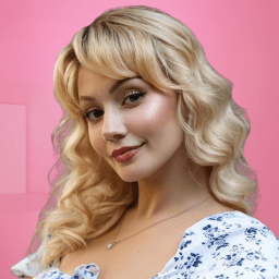 Woman with blonde curly hair, wearing a floral dress, smiling against a pink background.