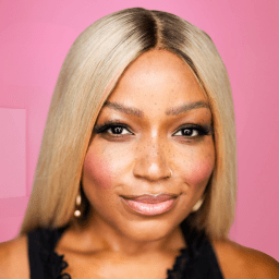 Smiling woman with long blonde hair, wearing pearl earrings, against a pink background.