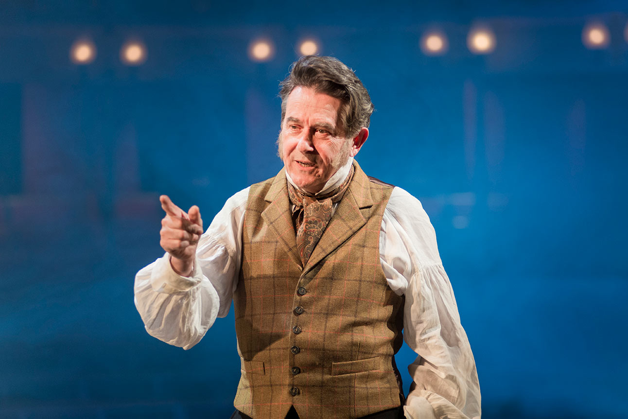Adrian Lukis as Mr Wickham, wearing a period costume, stands on stage with a thoughtful expression, pointing..