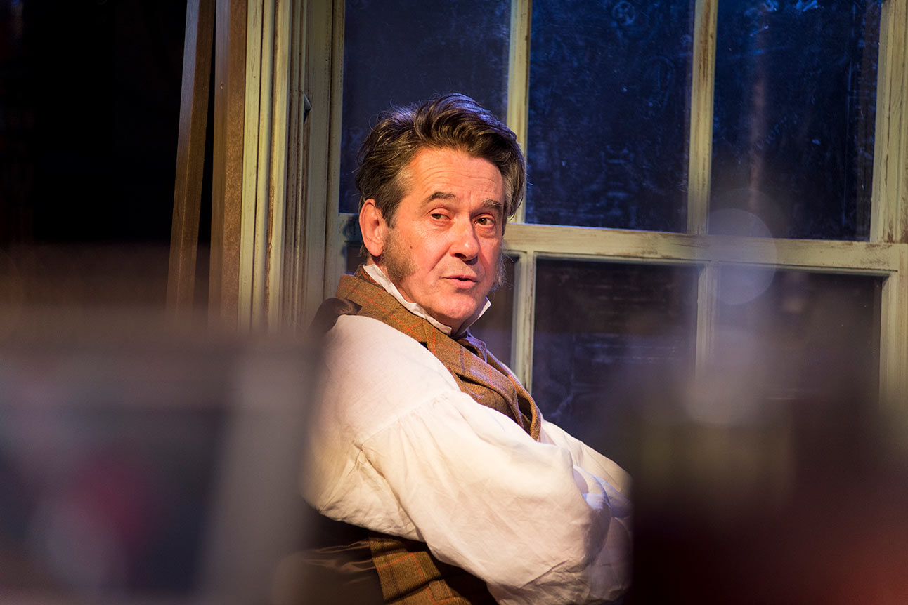 Adrian Lukis as Mr Wickham, in period costume, standing on stage next to a window.