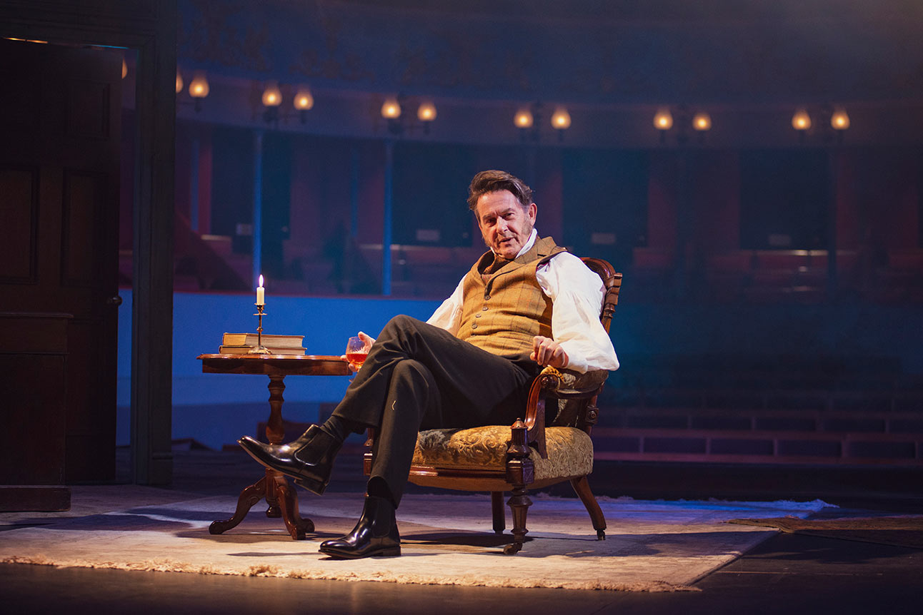 Adrian Lukis as Mr Wickham, dressed in period costume, sits on stage with a drink next to a table with a candle.