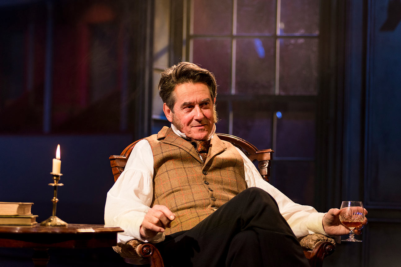 Adrian Lukis as Mr Wickham, dressed in period costume, sits on stage with a drink.