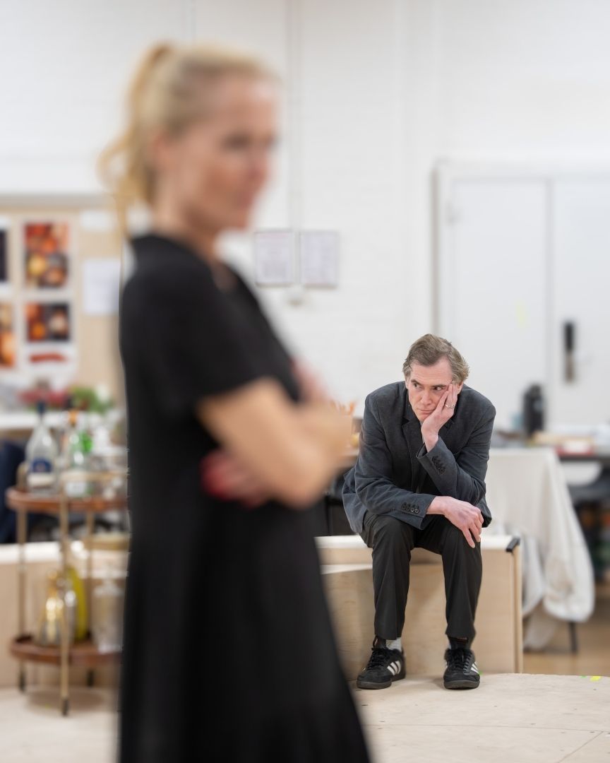 Billy Crudup sits thoughtfully on a bench, blurred woman in foreground, rehearsal room setting.