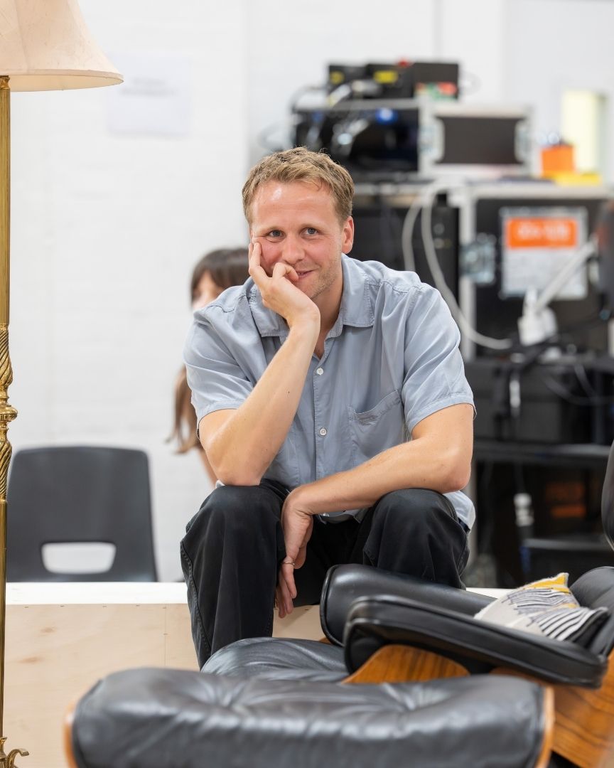 Josh Dylan in light blue shirt sitting, smiling thoughtfully, with hand on chin