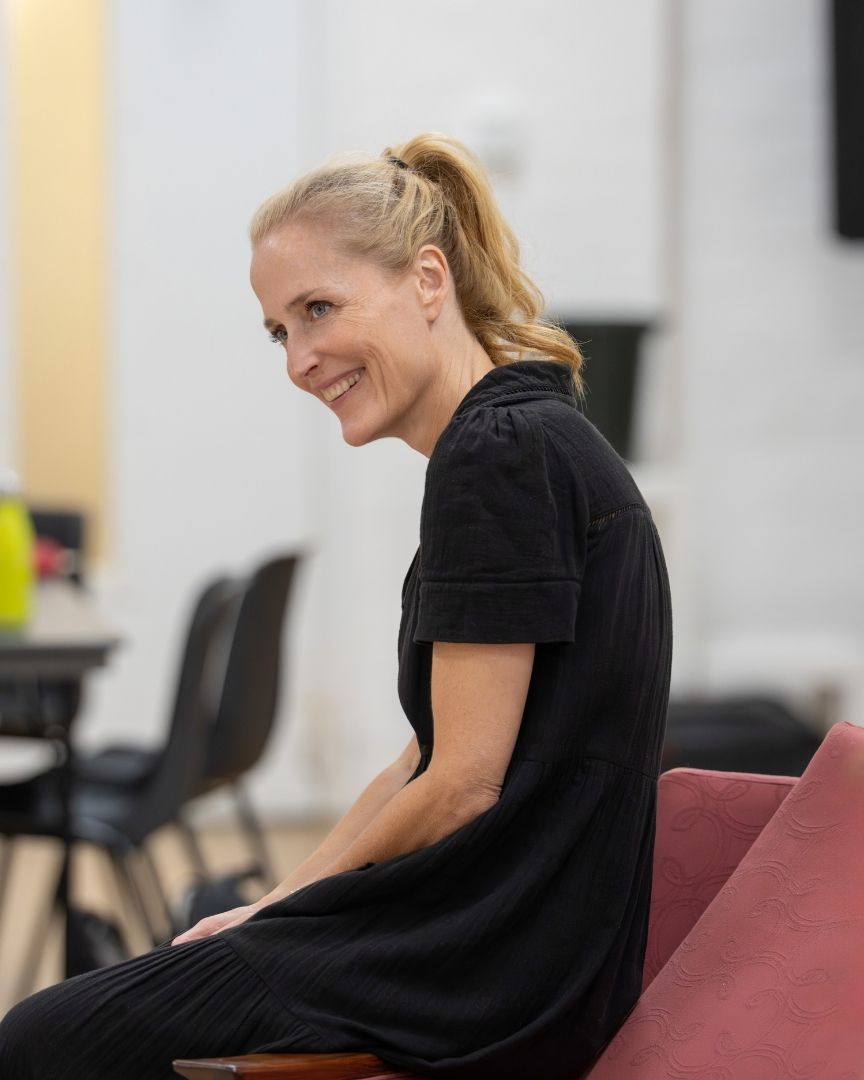 Gillian Anderson in a black dress sits smiling, with chairs and tables blurred in the background.