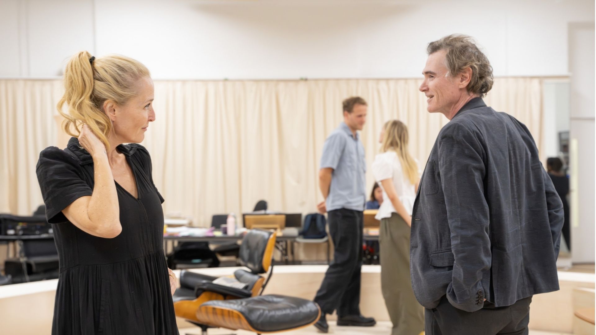 Two people converse in a rehearsal room; two others talk in the background, beige curtains visible.