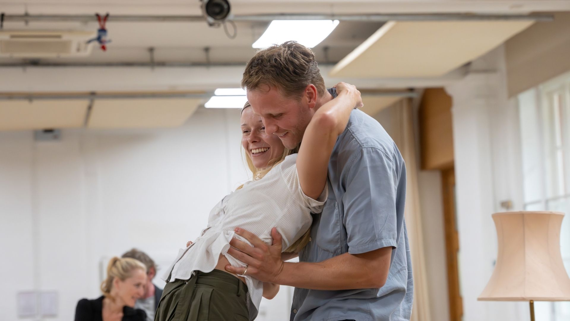 A man and woman embrace joyfully in a rehearsal room, both smiling, with bright overhead lighting.