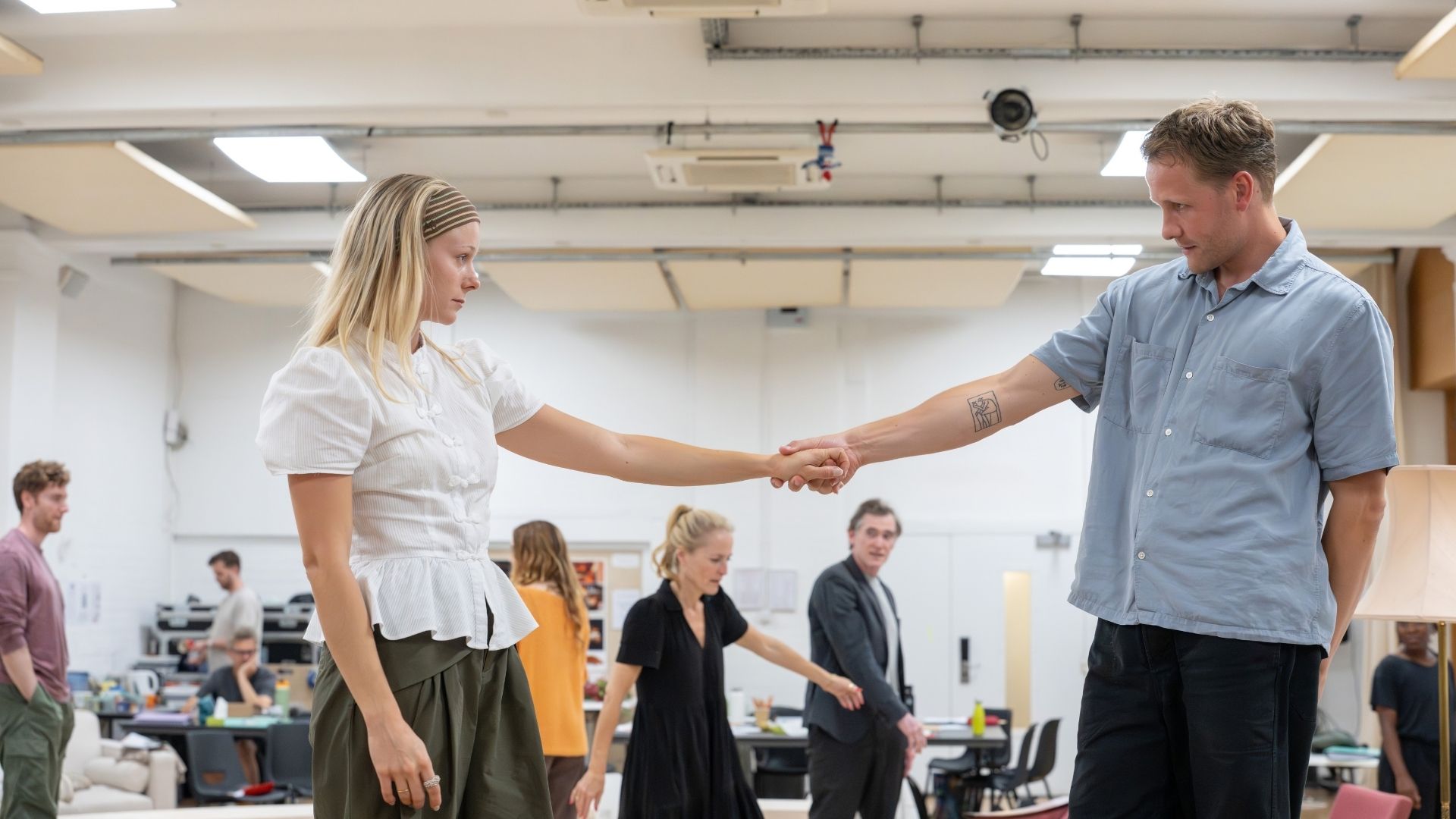 Two actors hold hands during rehearsal; others observe in the background