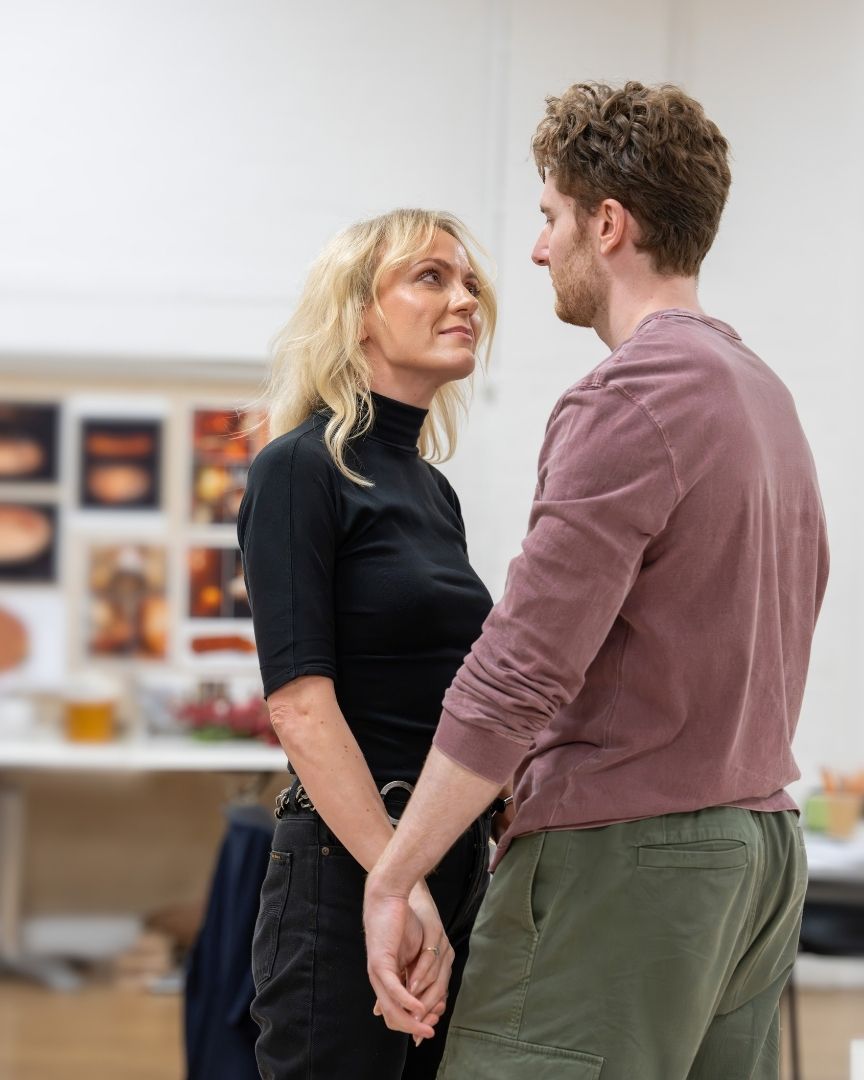 A man and woman hold hands, gazing at each other in a rehearsal room setting.