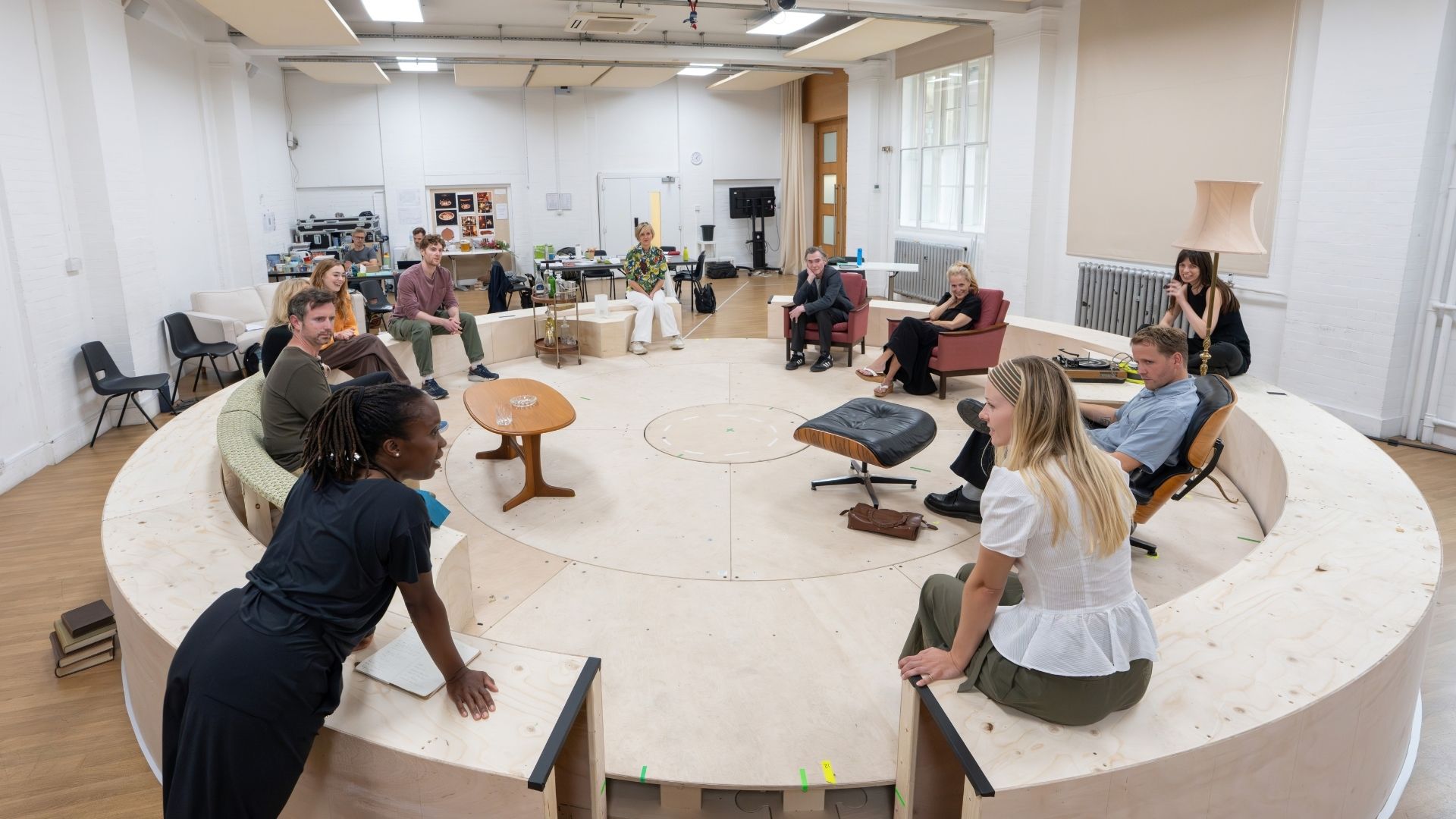 People seated in a circle on a stage set, with furniture and rehearsal props visible.