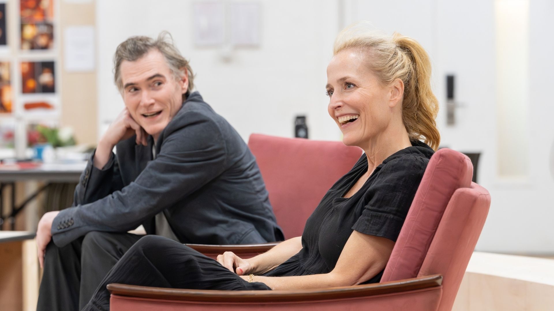 Gillian Anderson and Billy Crudup, seated on red chairs, laughing during rehearsal.