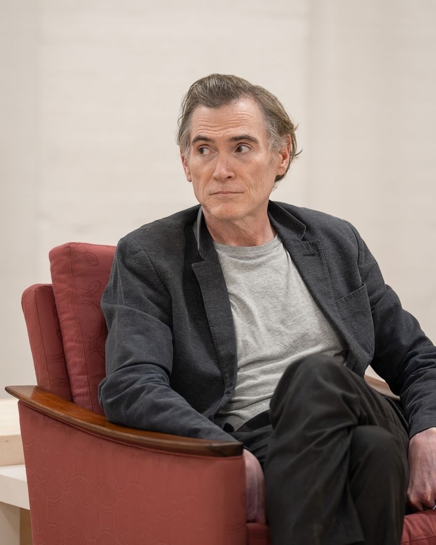 Billy Crudup in a grey shirt and dark blazer sits on a red chair, looking sideways.