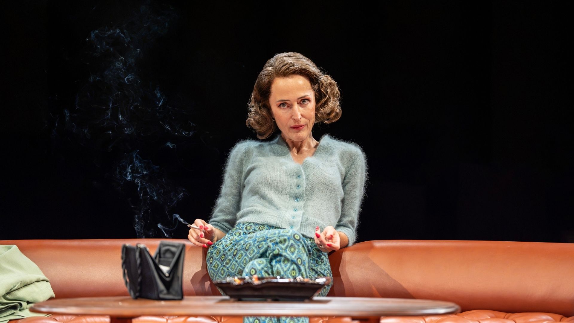A woman in a green cardigan and patterned skirt sits on a sofa, holding a cigarette.