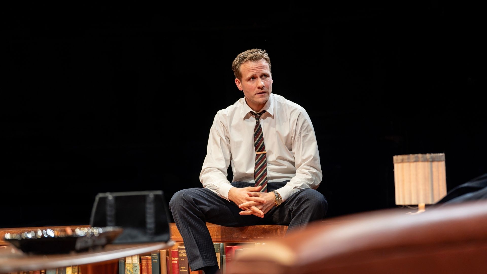 A man in a white shirt and tie sits on stage, surrounded by books.