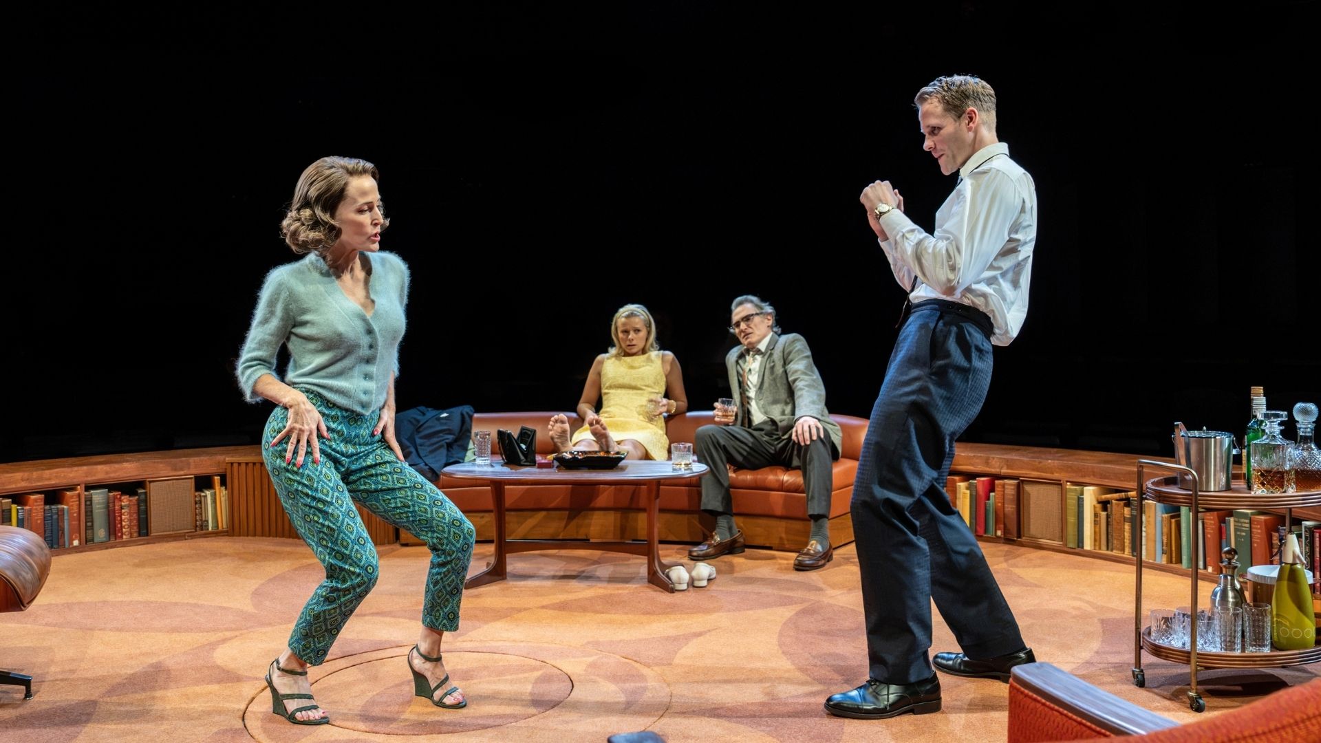 Two actors energetically dance in foreground; two seated actors watch from a sofa in background.