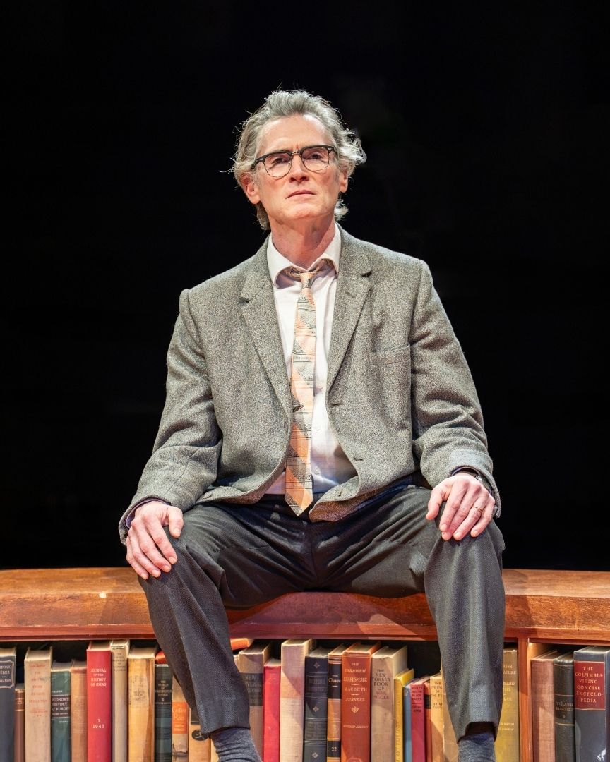 Man in grey suit and glasses sits on stage, surrounded by books, dark background.